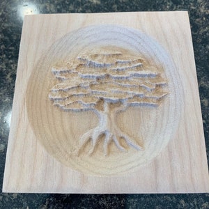 May include: A wooden plaque with a carved tree design. The tree has a thick trunk and branches with intricate details. The plaque is a light brown color and has a smooth finish.