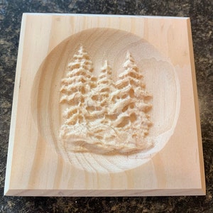 May include: A wooden plaque with a carved design of three pine trees in a circle. The plaque is light brown and has a smooth finish.