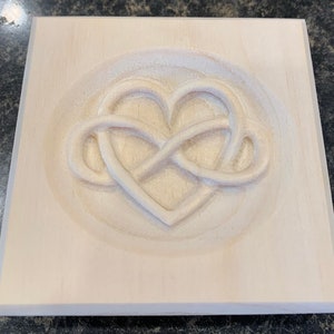 May include: A wooden plaque with a carved infinity symbol intertwined with a heart. The plaque is light brown and the carving is white.