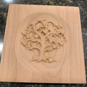 May include: A square wooden plaque with a carved tree design. The tree's leaves are heart-shaped. The wood has a light brown color and a visible grain.