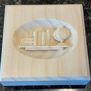 May include: A light-colored wooden plaque with a carved design. The design features a stack of books, a globe, and a shelf, all within an oval recess. The plaque is square with a light wood grain.