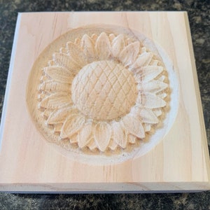 May include: A wooden plaque with a carved sunflower design. The sunflower is in the center of the plaque and is surrounded by a circle.