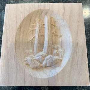 May include: A wooden plaque with a carved image of two trees in a forest setting. The trees are carved in relief, and the background is a smooth, light wood.