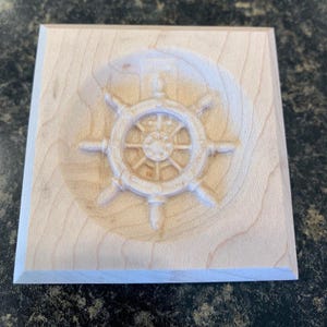 May include: A wooden square plaque with a carved wooden ship's wheel in the center.