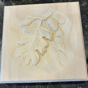 May include: A square light-colored wooden plaque featuring a relief carving of an oak leaf and two acorns centered within a circular recessed area. The natural wood grain is visible across the flat surface and the carved botanical design.
