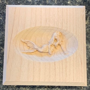 May include: A wooden plaque with a carved mermaid design. The mermaid is carved in relief and is lying on her side with her tail curled around her.