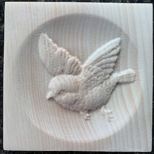 May include: A carved wooden square features a detailed relief of a bird in flight. The bird's wings are spread, and its feathers are intricately carved. The wood has a natural, light color, and the carving is set within a circular recessed area.