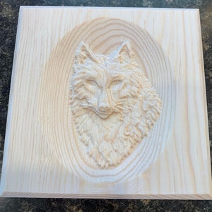 May include: A wooden plaque with a carved wolf head in the center. The wolf is carved in relief and has a detailed fur texture. The plaque is made of light wood and has a natural finish.