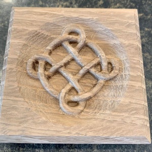 May include: A wooden square plaque with a carved Celtic knot design in the center. The knot is intricate and detailed, with a light brown color.