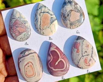 May include: A collection of six polished gemstone cabochons in various shapes. The stones display a range of colors, including cream, pink, gray, and burgundy, with unique veining and patterns. Each stone is displayed on a white surface.