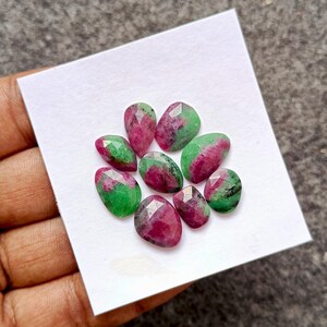 May include: A collection of eight faceted gemstones, each with a unique shape. The stones feature a vibrant combination of red and green hues, with a polished, reflective surface. The gemstones are arranged on a white surface.
