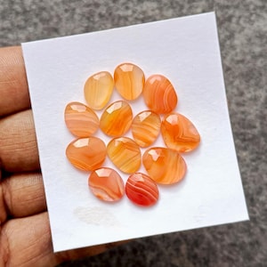 Op de afbeelding: Een verzameling ovaalvormige, gepolijste edelstenen in tinten oranje en rood. De stenen hebben een gestreept patroon en zijn gerangschikt op een wit oppervlak. De edelstenen worden waarschijnlijk gebruikt voor het maken van sieraden of verzamelen.