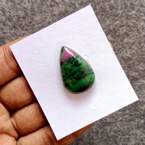 Puede incluir: Una gema pulida en forma de lágrima. Presenta una llamativa combinación de colores: verde intenso, negro y un rosa vivo en la parte superior. La gema se presenta sobre un fondo blanco.