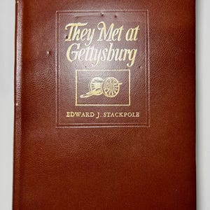 May include: A brown hardcover book titled "They Met at Gettysburg" by Edward J. Stackpole. The title and author's name are in gold lettering. The book cover features an embossed design of a cannon.