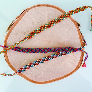 May include: Three colourful friendship bracelets with woven patterns rest on a wooden slice. The bracelets feature a variety of colours, including red, orange, yellow, green, blue, and purple. Each bracelet has a braided end.