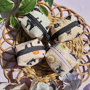May include: Four small zippered pouches with different patterns. The pouches are made of fabric and have a black zipper. The patterns include black cats, a pumpkin, a sun, and a butterfly.