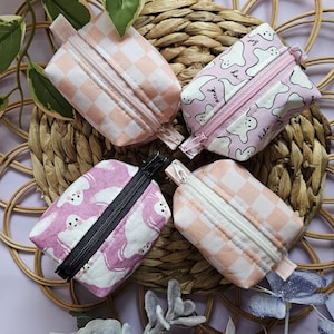 May include: Four small pink and white zippered pouches. Two pouches have a checkered pattern, and two pouches have a ghost pattern with the text "boo" and "seek" on them.