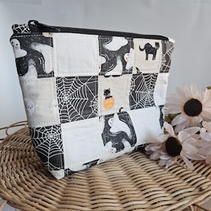 May include: A handmade Halloween-themed zippered pouch. The patchwork design features black and white squares with ghost, spiderweb, and black cat motifs. The bag is made of fabric and has a black zipper.
