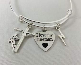 May include: Silver-toned adjustable bangle bracelet with three charms. Charms include a lineman on a utility pole, a heart-shaped charm with "I love my lineman" and a black heart, and a lightning bolt.