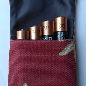 May include: A burgundy fabric pouch with a white floral pattern, holding four copper-colored batteries. The pouch has a black interior and a white trim at the top. The batteries are labeled with text and symbols.