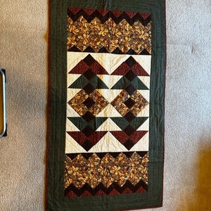 Puede incluir: Una colcha verde, marrón y roja con un patrón geométrico. La colcha está hecha de tela con un estampado de hojas y un patrón de chevron.