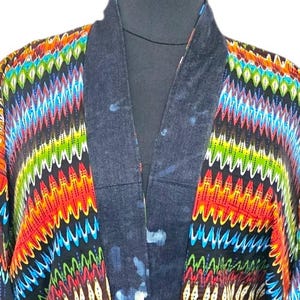 Op de afbeelding: Een kort jasje in kimonostijl met een levendig, veelkleurig zigzagpatroon in tinten rood, oranje, geel, groen en blauw. Het kledingstuk heeft een donkerblauwe bies met een gespikkeld effect langs de kraag, de manchetten en een lang strikceintuur.
