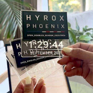 May include: Clear acrylic plaques with the text "HYROX PHOENIX" and event details. One plaque displays "SEPTEMBER 2025" and a time. Another shows "APRIL 2026". The plaques are held in front of a wooden frame.