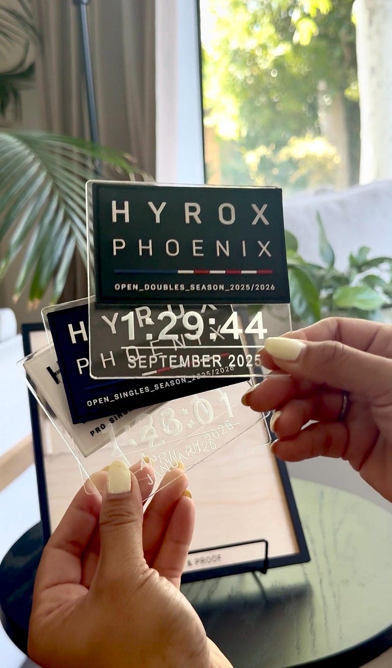 May include: Clear acrylic plaques with the text "HYROX PHOENIX" and event details. One plaque displays "SEPTEMBER 2025" and a time. Another shows "APRIL 2026". The plaques are held in front of a wooden frame.