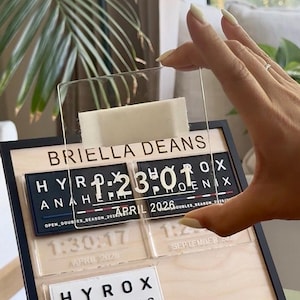 May include: A wooden desk calendar with a clear acrylic overlay. The calendar displays the name "BRIELLA DEANS" and event details, including "HYROX" and dates like "APRIL 2026". The time "1:23:01" is also visible. The calendar is held by a hand.