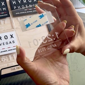 May include: A clear acrylic rectangle with the text "April Rose" and "1:53:0" printed on it. The rectangle has 3M PE foam tape on the back. The rectangle is being held in front of a black and white sign.
