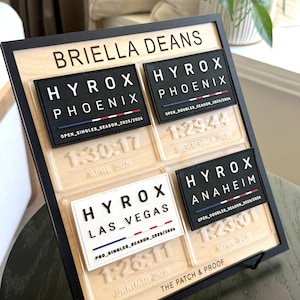 May include: A wooden plaque with a black frame displays four HYROX patches with event details. The patches are black and white with red, white, and blue stripes. The plaque also includes the name "BRIELLA DEANS" and the text "THE PATCH & PROOF".