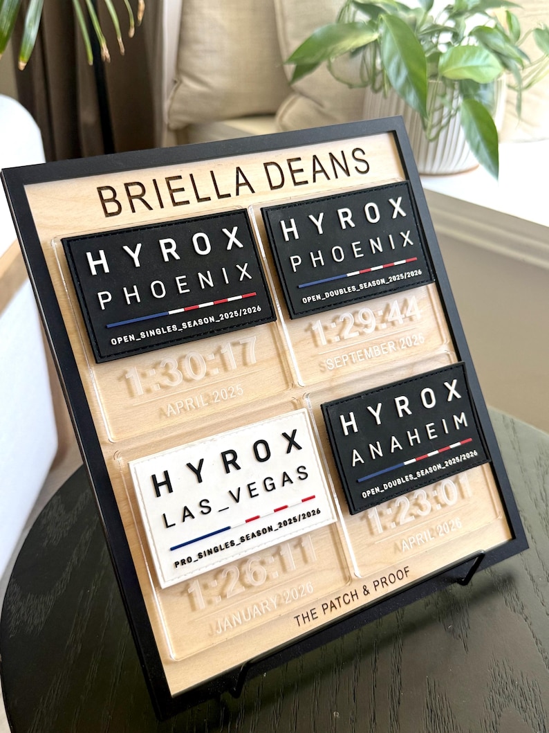 May include: A wooden plaque with a black frame displays four HYROX patches with event details. The patches are black and white with red, white, and blue stripes. The plaque also includes the name "BRIELLA DEANS" and the text "THE PATCH & PROOF".