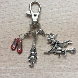 May include: A silver keychain with a charm of a witch flying on a broomstick, a charm of Dorothy from The Wizard of Oz, and two red ruby slipper charms.