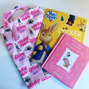 May include: A pink tote bag with a white and pink pattern featuring bunnies and the text "Mischief Maker hopping don't stopping". The bag is next to a Peter Rabbit sticker book and a pink book titled "The Tale of Jemima Puddle-Duck" by Beatrix Potter.