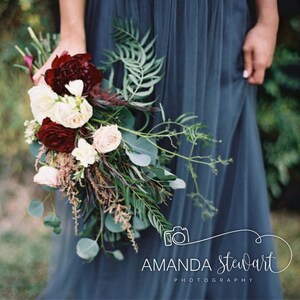 May include: A bride holds a bouquet of white and burgundy flowers with greenery. The bouquet is held in front of a blue bridesmaid dress. The image includes the text "Amanda Stewart Photography".