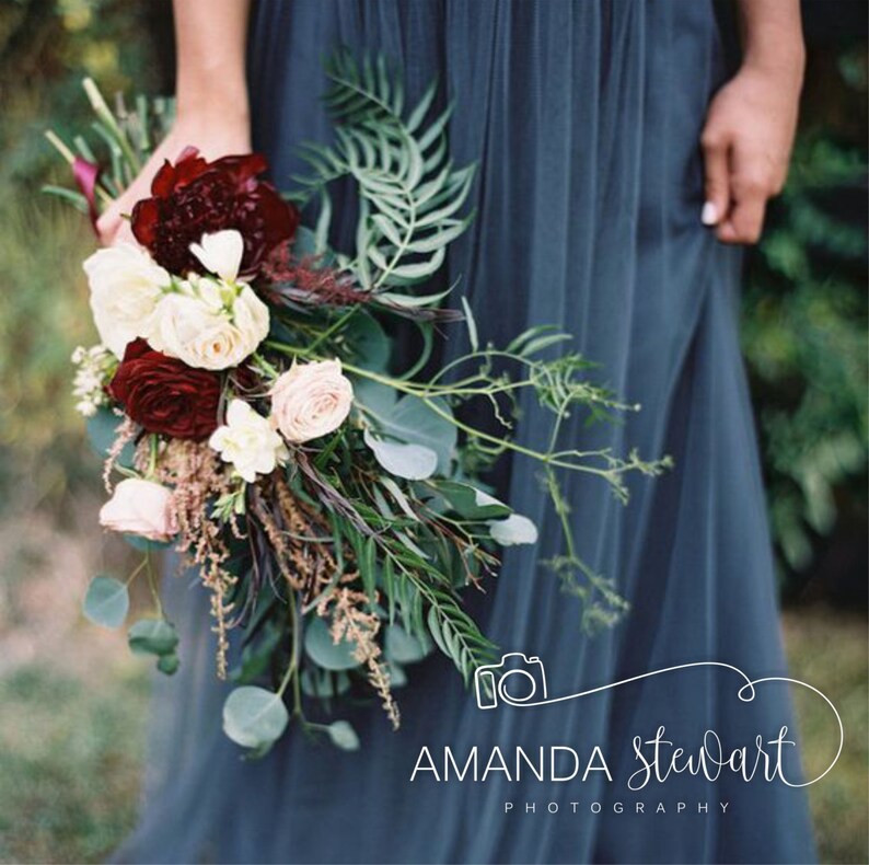 May include: A bride holds a bouquet of white and burgundy flowers with greenery. The bouquet is held in front of a blue bridesmaid dress. The image includes the text "Amanda Stewart Photography".