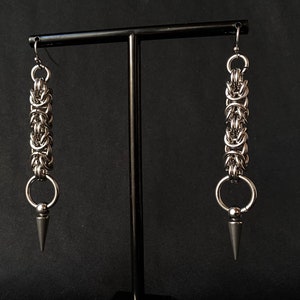 May include: A pair of silver earrings with a chain link design and a pointed tip. The earrings are hanging from a black stand.