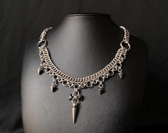May include: Silver-toned chainmail necklace with a gothic aesthetic. The necklace features a series of chain links and dangling cone-shaped spikes. A central pendant with a cross-like design and a longer spike adds to the edgy style. The necklace is displayed on a black mannequin.