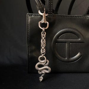 Può includere: Un portachiavi a forma di serpente argentato con un design a catena. Il portachiavi è attaccato a una borsa a mano in pelle nera con un grande logo circolare.