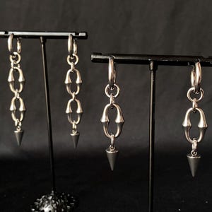 Puede incluir: Dos pares de pendientes de plata con un diseño de cadena y una punta puntiaguda. Los pendientes se muestran en un soporte negro.