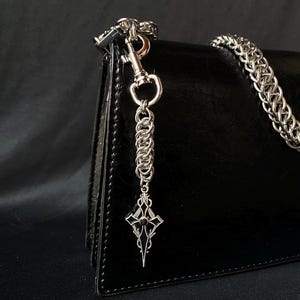 Könnte beinhalten: Eine schwarze Ledertasche mit einer silbernen Kette und einem silbernen Kreuzanhänger. Die Kette ist mit einem silbernen Verschluss an der Tasche befestigt.