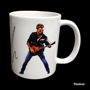 May include: White ceramic mug featuring a musician playing a guitar. The image shows the musician wearing sunglasses, a black jacket, and blue jeans. The mug has a curved handle and a signature on the side.