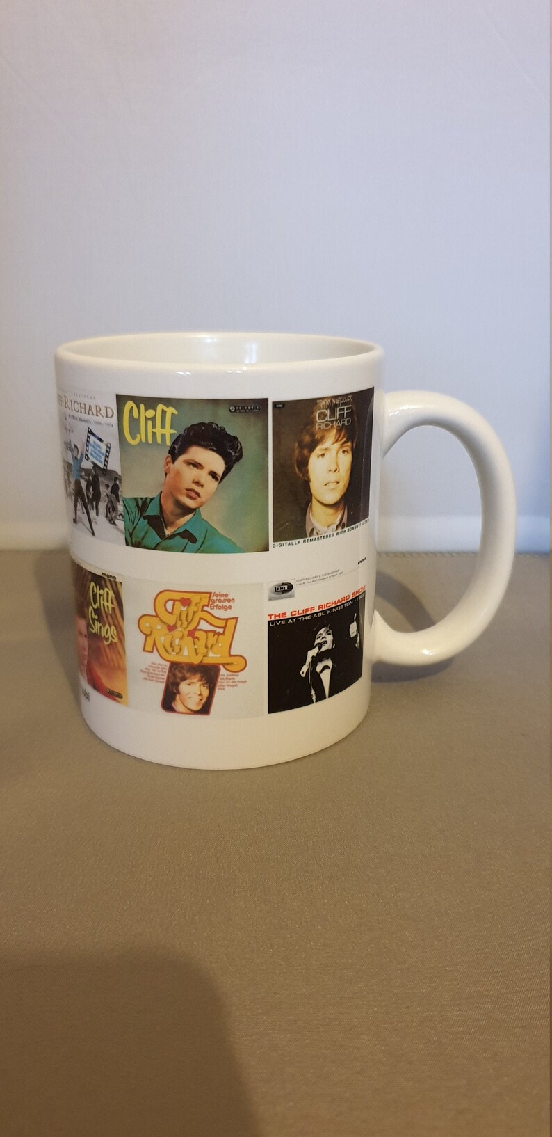 May include: White ceramic mug with a collage of Cliff Richard album covers. The covers feature various images of Cliff Richard, including a close-up of his face, a photo of him singing, and a photo of him performing on stage. The text on the covers includes "Cliff", "Cliff Richard", "The Cliff Richard Show", and "Live at the ABC, Kingston".