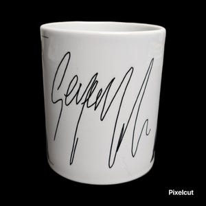 May include: White ceramic mug with a black handwritten signature design. The mug has a cylindrical shape and a smooth, glossy finish. The black signature is the primary design element, adding a unique touch to the mug.