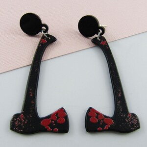 May include: A pair of dangle earrings shaped like axes. Each earring features a black axe head and handle with red paint splatters resembling blood. The earrings are attached to a circular black stud top with a silver-toned metal jump ring connector.