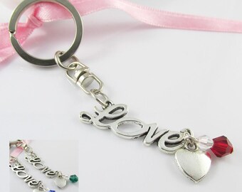 May include: A silver-toned metal keychain featuring a script-style word Love charm, a small heart charm, and a faceted red crystal bead. The keychain includes a circular ring attached to a light pink ribbon and a lobster clasp connector.