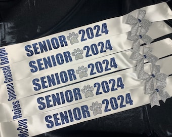 May include: Five white satin sashes featuring the dark blue glitter text SENIOR 2024 with a silver glitter paw print icon. Each sash includes a silver glitter bow accent on the right side and personalized names printed vertically in blue.