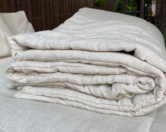 May include: A folded, natural-colored linen quilt and pillow set. The quilt is thick and quilted with a subtle pattern. The neutral color and texture suggest a focus on comfort and natural materials. The set is neatly arranged, suggesting a focus on home decor.
