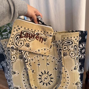 May include: A quilted tote bag and a matching zippered pouch featuring a tan and black paisley bandana print. The pouch is embroidered with the name Lauren in dark brown thread. Both items have a structured, padded design with fabric handles.