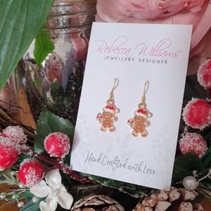 May include: Gold-tone gingerbread man earrings with red and white accents. The earrings are displayed on a white card with the text "Rebecca Williams JEWELLERY DESIGNER" and "Hand Crafted with Love". Festive holiday jewelry.